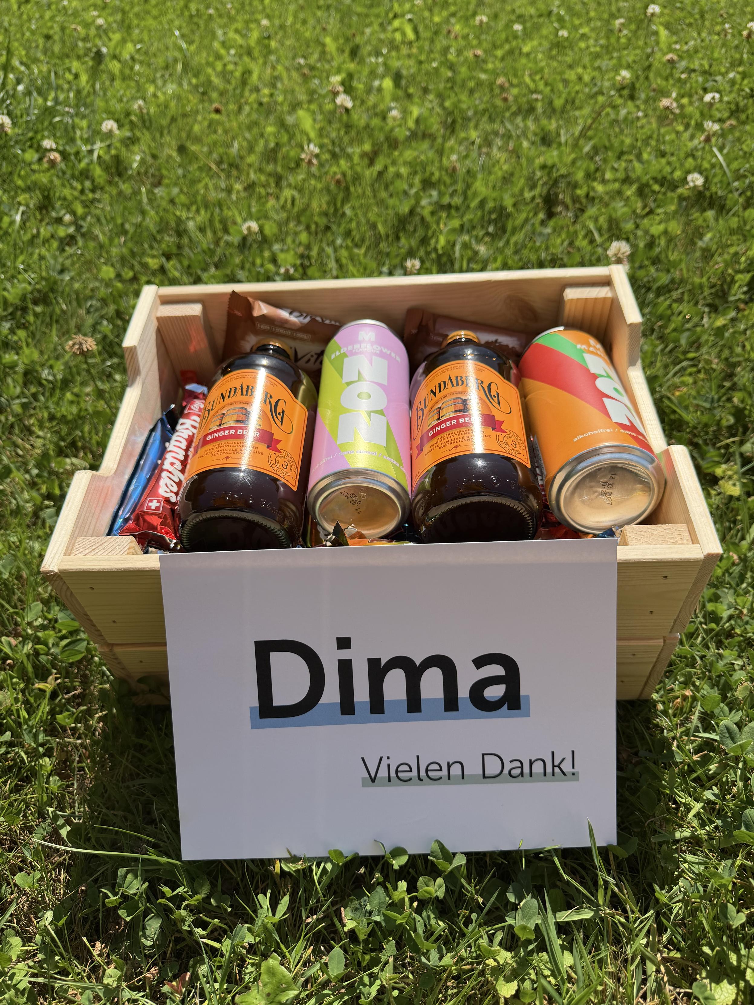 A wooden crate sitting in grass, filled with two bottles of Bundaberg ginger beer, two canned drinks (one pink, one orange), and a couple of chocolate bars. A white sign propped against the crate reads 'Dima' in large bold letters, with 'Vielen Dank!' written below.