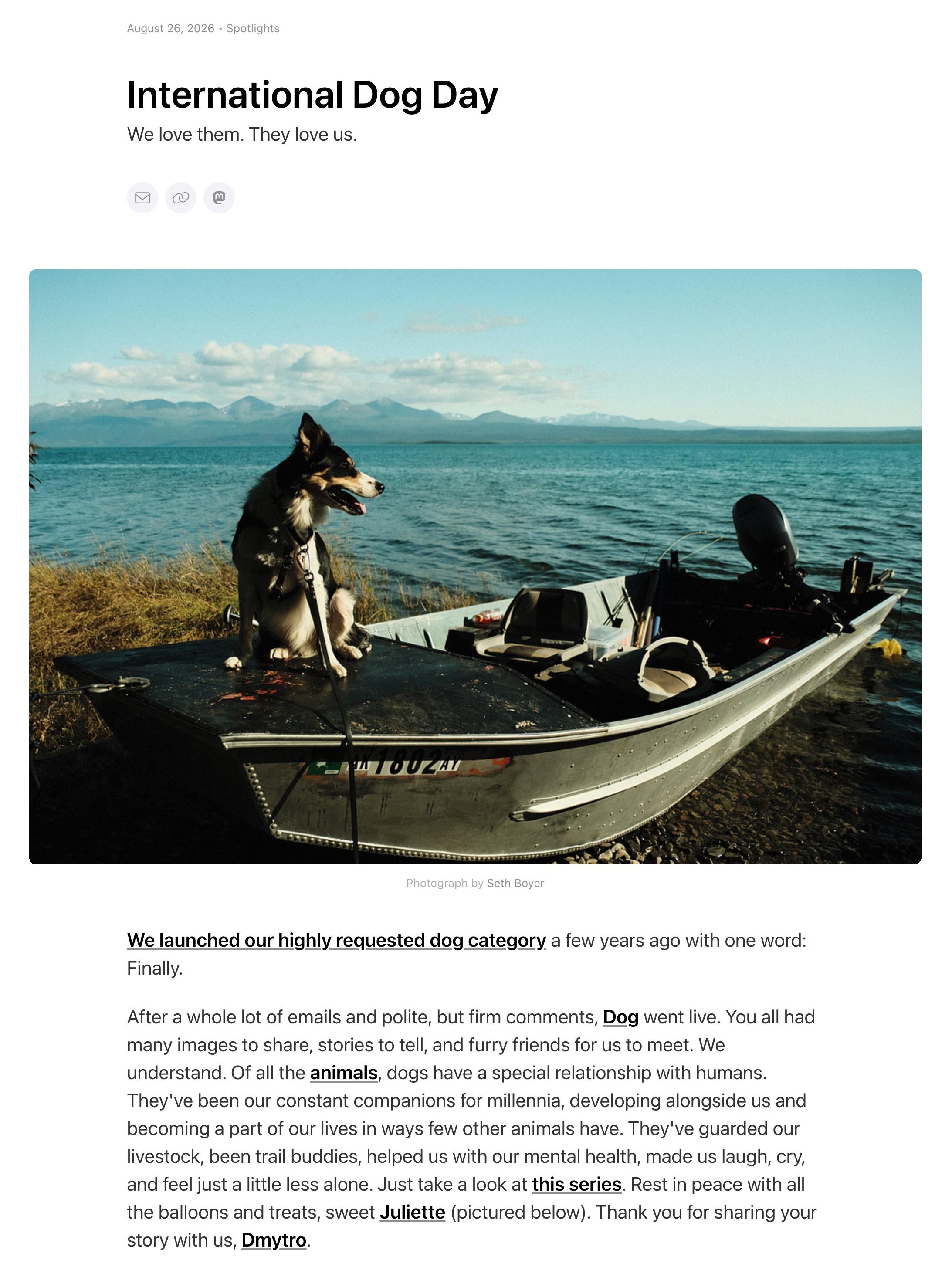 Screenshot of a glass.photo Spotlight page dated August 26, 2026. Large heading "International Dog Day" and the line "We love them. They love us." Three small round buttons for mail, link and Mastodon sit under the heading. The main photograph, credited "Photograph by Seth Boyer", shows a black, white and tan dog on a leash sitting on the front deck of a silver aluminium fishing boat pulled onto a pebbly lake shore, mountains and a blue sky behind it. Boat registration readable as AK 1802AY. Body text begins "We launched our highly requested dog category a few years ago with one word: Finally." Later lines name Juliette and Dmytro: "Rest in peace with all the balloons and treats, sweet Juliette (pictured below). Thank you for sharing your story with us, Dmytro.